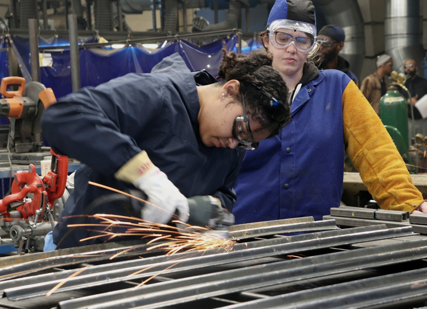 A trainee grinding steel while an instructor looks on